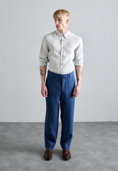 Camisa de lino gris claro con botones, combinada con pantalones azules ajustados que presentan un detalle de cinturón lateral y zapatos de cuero marrón.
