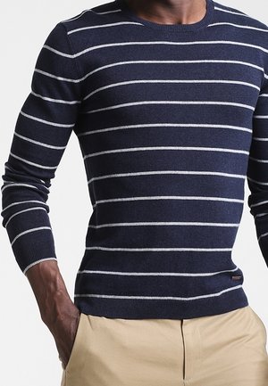Mann trägt einen dunkelblauen Langarmpullover mit dünnen weißen horizontalen Streifen und beige Hosen, Hände in den Taschen.