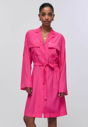 Kobieta ubrana w jasnoróżową sukienkę koszulową z długim rękawem, paskiem i kieszeniami na biuście, złote kolczyki koła oraz wielowarstwowe złote naszyjniki, stojąca na białym tle.