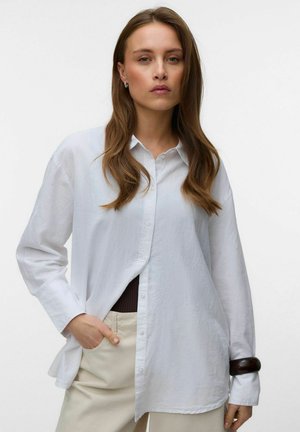 Žena s dlhými hnedými vlasmi nosí nadmernú bielu košeľu na gombíky, béžové nohavice, tmavý náramok a má jednu ruku v vrecku na bielom pozadí.