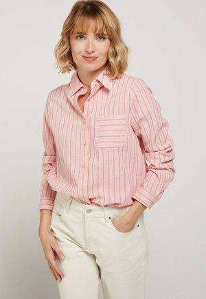 Femme aux cheveux courts et ondulés portant un chemisier à manches longues rayé rose et blanc, rentré dans un pantalon taille haute écru, main dans la poche.