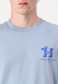 Lys blå bomull t-skjorte med rund hals. Har et blått grafisk trykk med teksten "HUGO" og "solarsphere tour 1998" på forsiden.