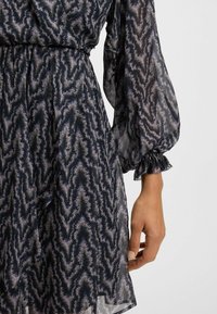 Abito a maniche lunghe, trasparente, di colore blu navy con un motivo astratto ondulato in diverse tonalità di grigio. Presenta polsini arricciati e una vita sagomata.