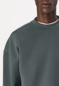 Mörkgrön sweatshirt i mjukt tyg, med rundad halsringning, ribbade ärmslut och sömnadsdetaljer längs axlarna.