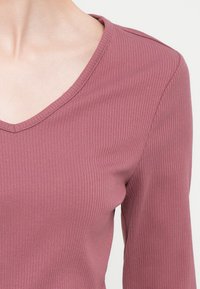 Αυτοκόλλητη πλεκτή μπλούζα σε απαλό ροζ χρώμα, με V-λαιμόκοψη και μακριά μανίκια. Το ύφασμα έχει μαλακή υφή με ελαφριά ελαστικότητα.