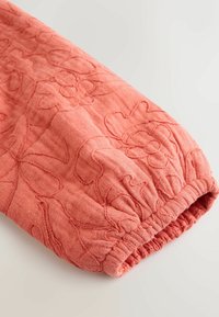 Korallszínű steppelt szövet ujj, virágmintás hímzéssel és ráncolt elasztikus mandzsettával.