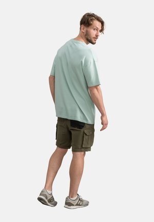 Helles teal Baumwoll-T-Shirt mit lockerer Passform, kombiniert mit olivgrünen Cargo-Shorts mit Taschen. Trägt graue Sportschuhe.