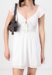 Vit kortärmad klänning med V-ringning och dragsko detaljer vid midjan. Texturen ser lätt ut; parat med en liten mörk axelväska.