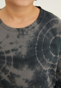 Sivá dlhá tričko s dlhým rukávom vyrobené z bavlny, s kruhovým tie-dye vzorom v tmavších a svetlejších odtieňoch a s okrúhlym výstrihom.