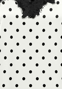 Tissu blanc avec des pois noirs uniformément espacés et un détail de bordure en dentelle noire le long du bord supérieur.