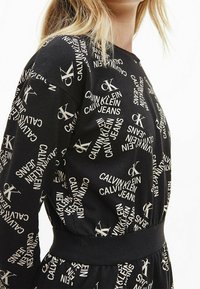 Musta kangastoppi, jossa on hajallaan valkoista "CALVIN KLEIN JEANS" -tekstiä. Suunnittelussa on pyöreä kaula-aukko ja tiukka vyötärö.