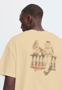 Crèmekleurig katoenen T-shirt met een geprinte afbeelding op de achterkant met twee figuren en de tekst "la dolce vita" in rode schrijfletters.