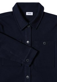 Chaqueta azul marino con cuello tipo solapa, dos bolsillos frontales y botones a presión plateados. Tejido texturizado con un sutil patrón de espiga.