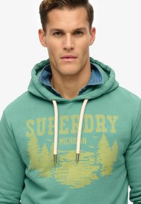 Sudadera verde con un gráfico amarillo de árboles y un lago. Tiene una capucha con cordones y una camisa de mezclilla de varias capas debajo.