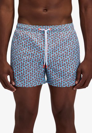 Hombre que lleva pantalones cortos de baño azul claro con un patrón repetido de pequeños trozos de sandía roja y un cordón blanco en la cintura.