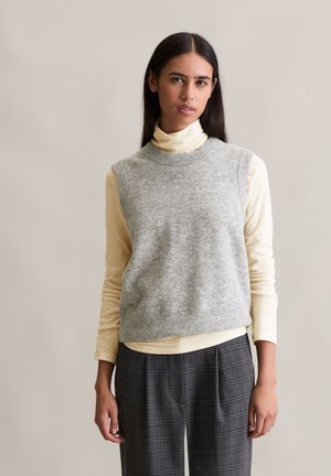 Grijze mouwloze trui over een lichtgele turtleneck, gecombineerd met grijze geruite broek. Zachte stof, casual ontwerp, met een relaxed fit.