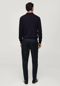 Mężczyzna w czarnym swetrze, ciemnych dopasowanych spodniach i czarnych eleganckich butach stoi przodem do jednolitego jasnoszarego tła.