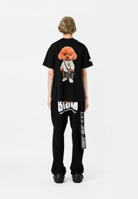 Camiseta negra con gráfico de perro de dibujos animados de gran tamaño en la parte posterior, combinada con pantalones negros que presentan texto y patrones en blanco llamativos. Tejido texturizado.
