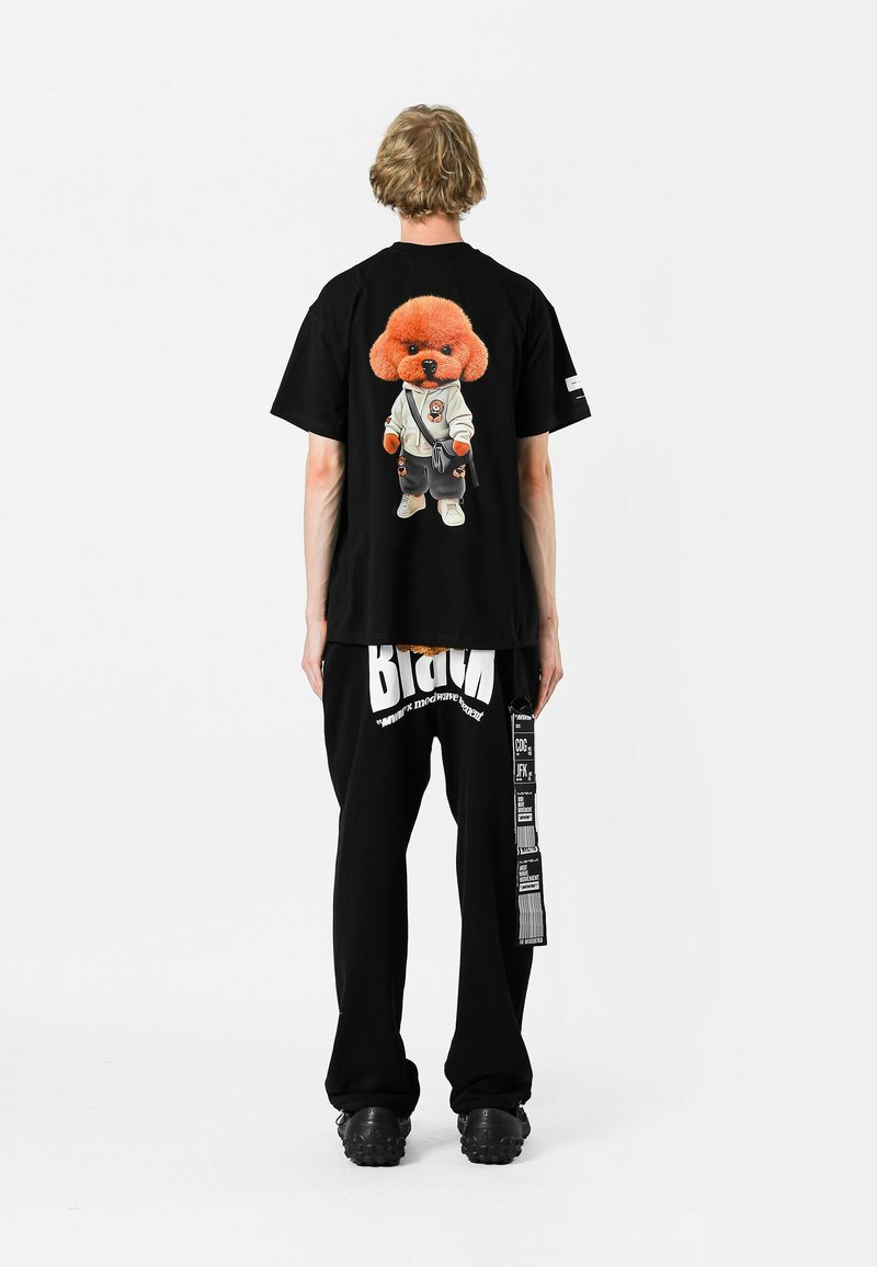 Camiseta negra con gráfico de perro de dibujos animados de gran tamaño en la parte posterior, combinada con pantalones negros que presentan texto y patrones en blanco llamativos. Tejido texturizado.
