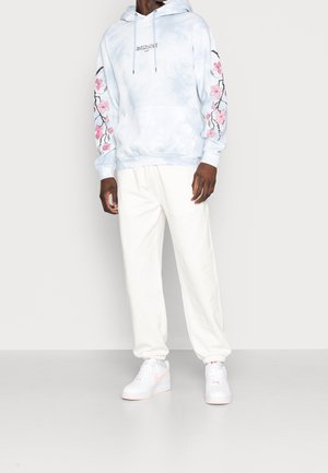 Hoodie tie-dye bleu clair avec broderie florale rose sur les manches, associé à un pantalon de survêtement blanc et des baskets blanches avec des accents roses.