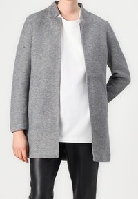 Cappotto grigio in misto lana con un design minimalista, colletto alto e maniche lunghe, indossato sopra una camicia bianca e pantaloni neri.