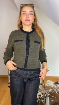 Cardigan de lã verde com detalhes em azul-marinho, fechamento com botões, duas bolsos frontais, combinado com jeans de denim escuro e uma bolsa metálica.