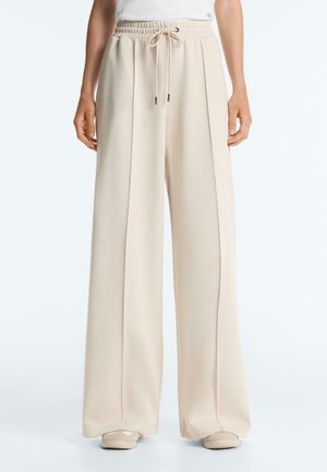 Person wearing high-waisted, wide-leg beige trousers with front pleats and drawstring waist, paired with beige shoes.