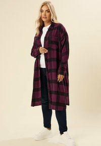 Camicia lunga a scacchi viola e nera, realizzata in tessuto morbido, con apertura a bottoni, taschino sul petto e vestibilità rilassata. Indossata sopra una maglietta bianca.