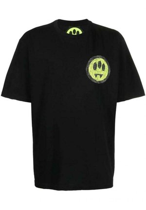 Mężczyzna w luźnej czarnej koszulce z małym żółto-zielonym emblematem z trzema czarnymi oczami i dwoma kłami na górnej lewej stronie klatki piersiowej.