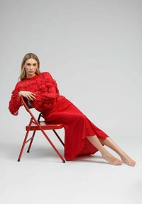 Top en dentelle rouge avec des accents volants associé à une longue jupe rouge élégante. Modèle assis sur une chaise rouge, pieds nus, avec un fond neutre.