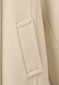 Beżowy wełniany płaszcz zbliżenie, prezentujący lekko teksturowaną tkaninę, charakterystyczną prostokątną naszywaną kieszeń oraz czyste detale szwów.