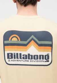 Κοντινό πλάνο της πλάτης ενός ατόμου που φοράει μπεζ πουκάμισο με λογότυπο σε ρετρό στυλ "Billabong Adventure Division" με γραφικό στοιχείο ήλιου και βουνού.