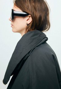 Czarny oversize płaszcz z wysokim kołnierzem, miękką teksturą, noszony z dużymi czarnymi okularami przeciwsłonecznymi i małą kolczykiem w kształcie kółka, widoczny z boku.