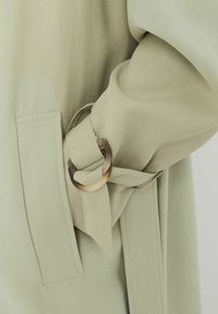 Veste ceinturée vert clair avec une texture lisse, dotée d'une boucle arrondie et translucide ainsi que de poches latérales. Détail de la ceinture et du tissu montré.