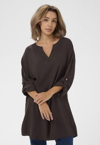 Tunic in lino marrone con scollatura a V, maniche tre quarti con dettagli a bottoni e vestibilità ampia, abbinato a jeans blu.