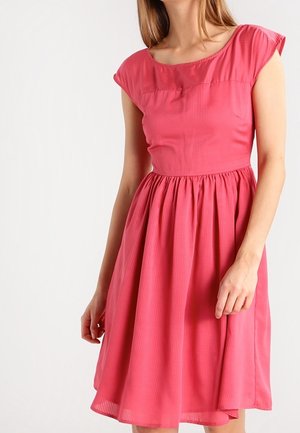 Žena ve zcela bezrukávových, kolenní délky, jasně růžových šatech s nařaseným pasem a texturovanou látkou, stojící na jednobarevném bílém pozadí.