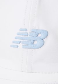 Hvit caps med en brodert lys blå New Balance-logo. Har synlige sømmer og en strukturert design. Myk, glatt stoff.