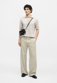 Kiharatukkainen mies, jolla on beige lyhythihainen neulepikeepaita, leveät beiget housut, mustat loafers-mokkanilkkurit ja musta olkalaukku.