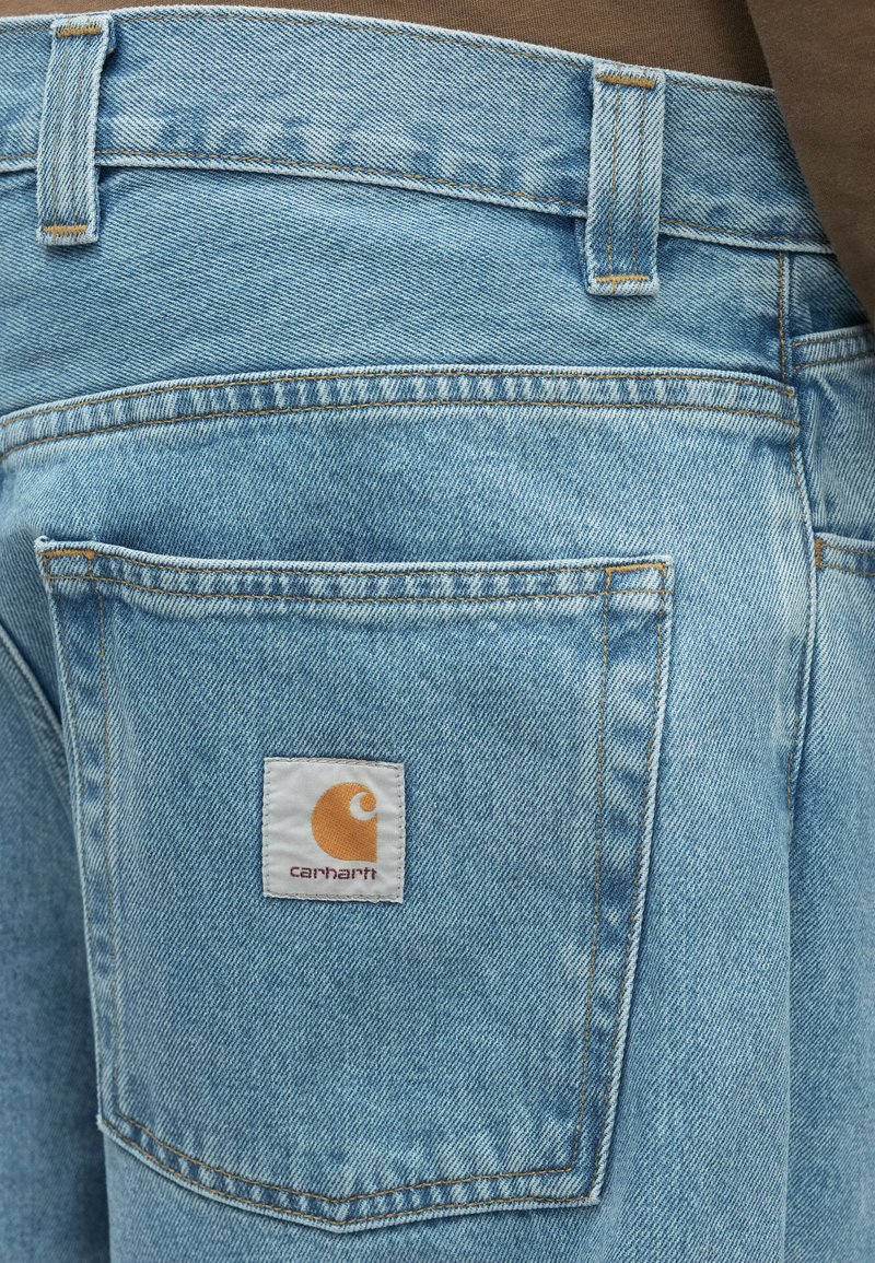 Pantalones vaqueros de mezclilla azul claro con un bolsillo trasero que presenta un parche blanco rectangular con un logo en naranja y texto en negro.
