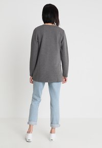 Szary top z długim rękawem o prostym kroju, w połączeniu z jasnoniebieskimi podwiniętymi jeansami i białymi wsuwanymi butami. Gładka faktura i luźne dopasowanie.