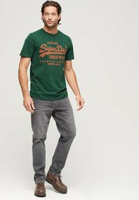 T-shirt in cotone verde con stampa grafica arancione in stile vintage. Abbinato a jeans grigi scuri e scarpe in pelle marrone.