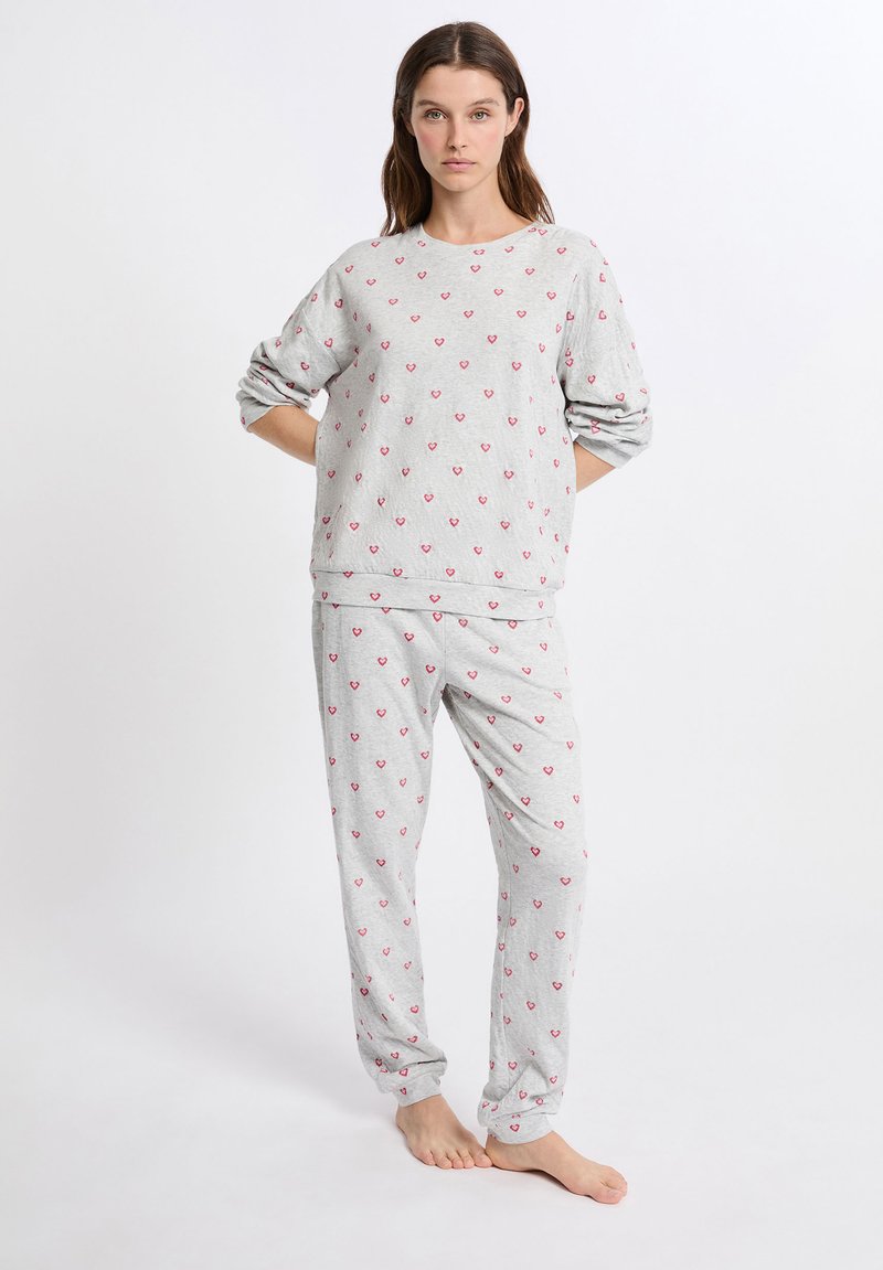 Conjunto de pijama gris que incluye un sudadera de cuello redondo y pantalones, ambos con pequeños patrones de corazones rosas. Tejido suave, corte relajado y puños en los tobillos.