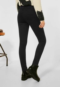 Musta kapea farkut korkealla vyötäröllä ja kahdella taka-taskulla, yhdistetty mustiin nilkkureihin. Kangas näyttää joustavalta ja sileältä.