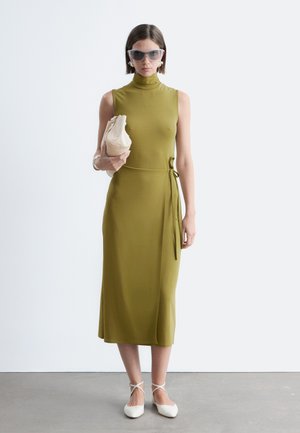 Femme portant une robe midi sans manches vert olive et des lunettes de soleil oversize, tenant une pochette beige, debout sur un sol gris contre un mur blanc.