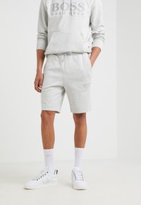 Homme portant un sweat à capuche Hugo Boss gris clair et un short assorti, avec des chaussettes blanches et des baskets blanches, se tenant devant un fond uni.
