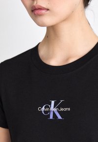 Nahaufnahme einer Person, die ein schwarzes T-Shirt mit einem blauen und weißen "Calvin Klein Jeans"-Logo auf der Brust trägt.