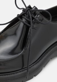 Black leather shoes with a glossy finish, round toe, and black laces, featuring a thick, textured rubber sole.