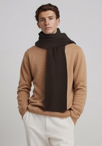 Écharpe côtelée marron drapée autour du cou, associée à un pull en maille beige. Le mannequin pose sur un fond gris clair.