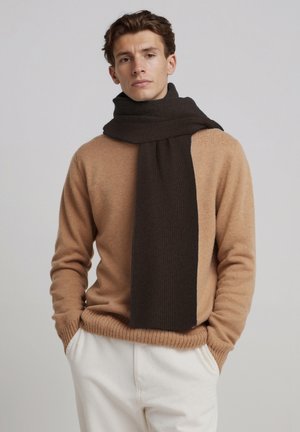 Écharpe côtelée marron drapée autour du cou, associée à un pull en maille beige. Le mannequin pose sur un fond gris clair.