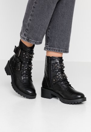 Personne portant des bottes de combat noires cloutées avec fermetures éclair latérales, boucles et cuir texturé, associées à un jean gris foncé raccourci.